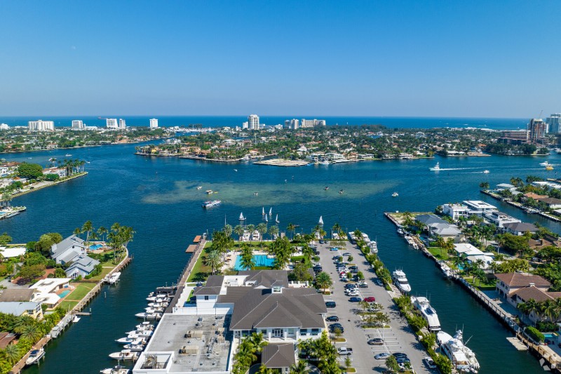 Aerial view of Harbor Beach, Fort Lauderdale, showcasing an upscale marina surrounded by luxury waterfront homes, calm blue waterways, and high-rise buildings in the distance along the coastline, under a clear blue sky.