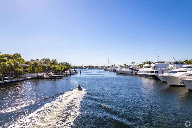 View of a calm waterway in Victoria Park, Fort Lauderdale, featuring luxury yachts docked on the right, waterfront homes on the left, and a jet skier creating a trail across the clear blue water under a sunny sky.