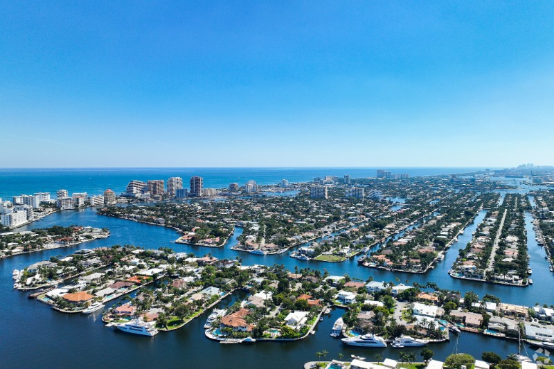 Aerial view of Sunrise Key in Fort Lauderdale, showcasing a series of waterfront homes and canals leading to the Atlantic Ocean, with high-rise buildings and the city skyline in the distance under a clear blue sky.