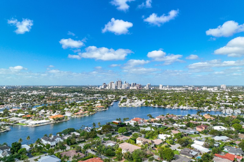 Scenic aerial view of Sunrise Intracoastal in Fort Lauderdale featuring luxury homes along the waterway, lush greenery, and a backdrop of the downtown skyline under a vibrant blue sky with scattered clouds.