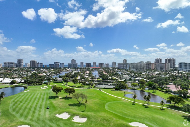 Aerial view of Coral Ridge Country Club in Fort Lauderdale, showcasing a lush golf course with sand traps and ponds, surrounded by palm trees, residential homes, and high-rise buildings under a partly cloudy sky.