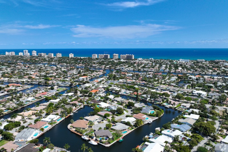 Aerial view of The Landings neighborhood in Fort Lauderdale, featuring winding canals lined with waterfront homes, lush greenery, and a backdrop of high-rise buildings and the Atlantic Ocean under a clear blue sky.