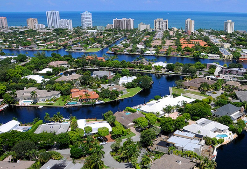 Aerial view of Bay Colony in Fort Lauderdale, showcasing a luxurious waterfront neighborhood with large homes, winding canals, abundant greenery, and a backdrop of high-rise condominiums along the Atlantic Ocean.