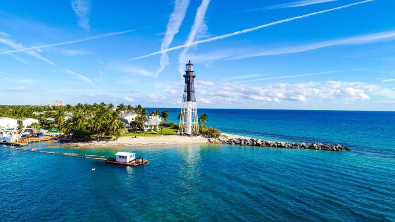 Aerial view of Lighthouse Point, Florida, featuring a historic black-and-white lighthouse on a sandy peninsula surrounded by clear turquoise water, with nearby palm trees, small buildings, and a floating dock under a bright blue sky with white contrails.