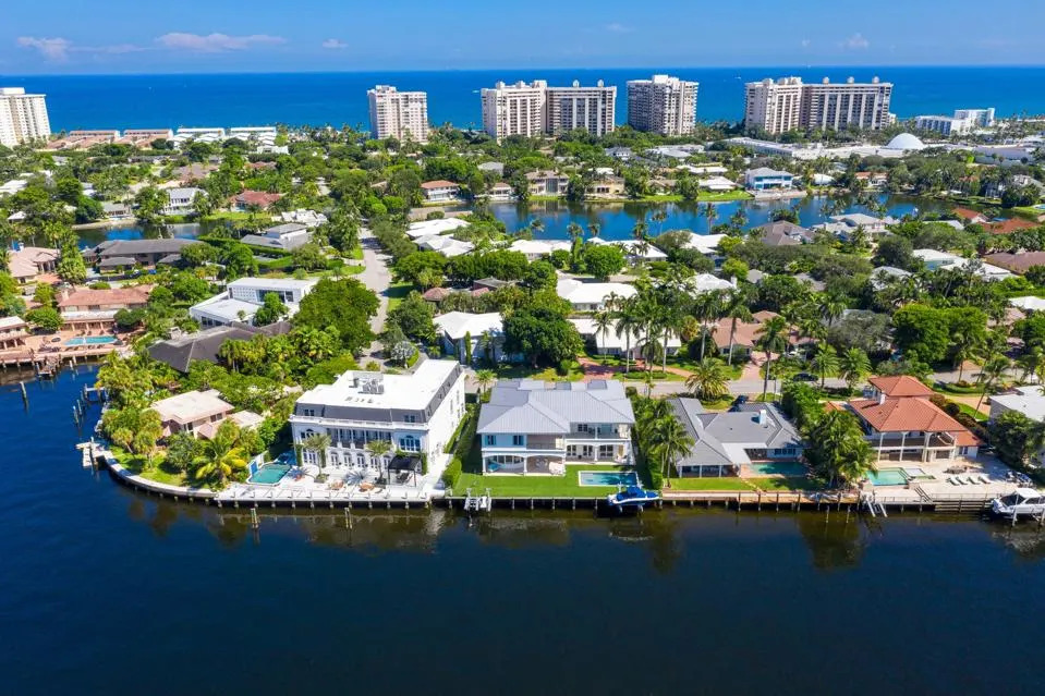 Aerial view of Sea Ranch Lakes, a waterfront residential community in Fort Lauderdale, featuring upscale homes with private docks along a calm canal, lush tropical landscaping, and a backdrop of tall oceanfront condos with the Atlantic Ocean in the distance.