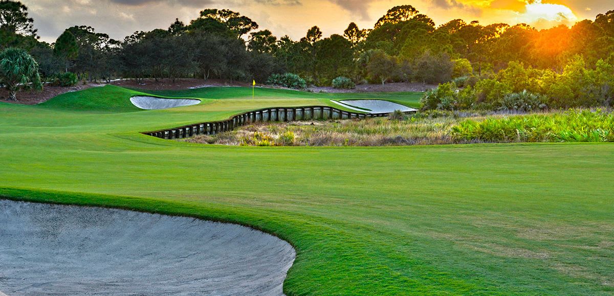 A scenic golf course at sunset, featuring lush green fairways, a sand trap, and a wooden bridge over a water hazard.
