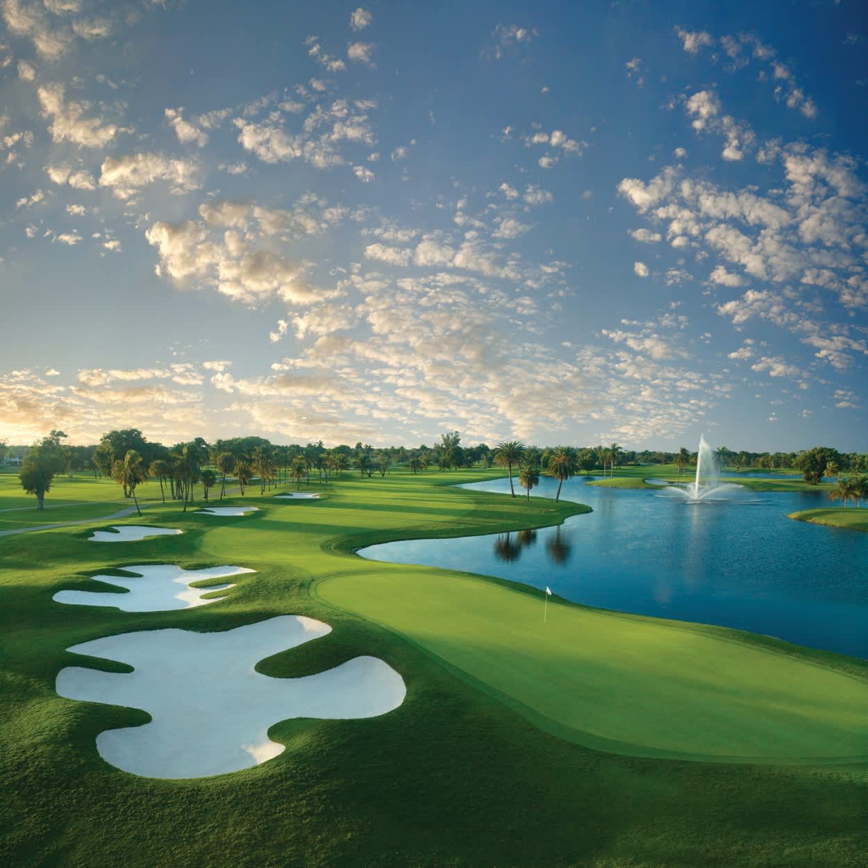 A scenic golf course with lush green fairways, sand traps, a pond, and a fountain under a blue sky with scattered clouds.