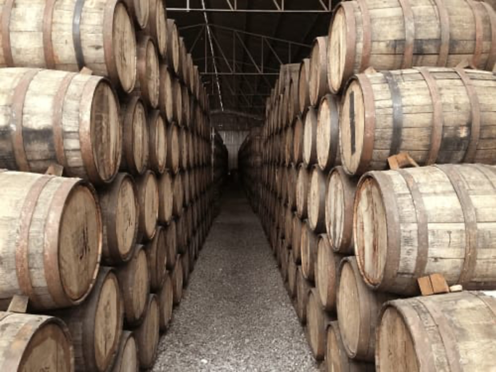 A long aisle lined with stacked wooden barrels under a roof, creating a rustic storage space.