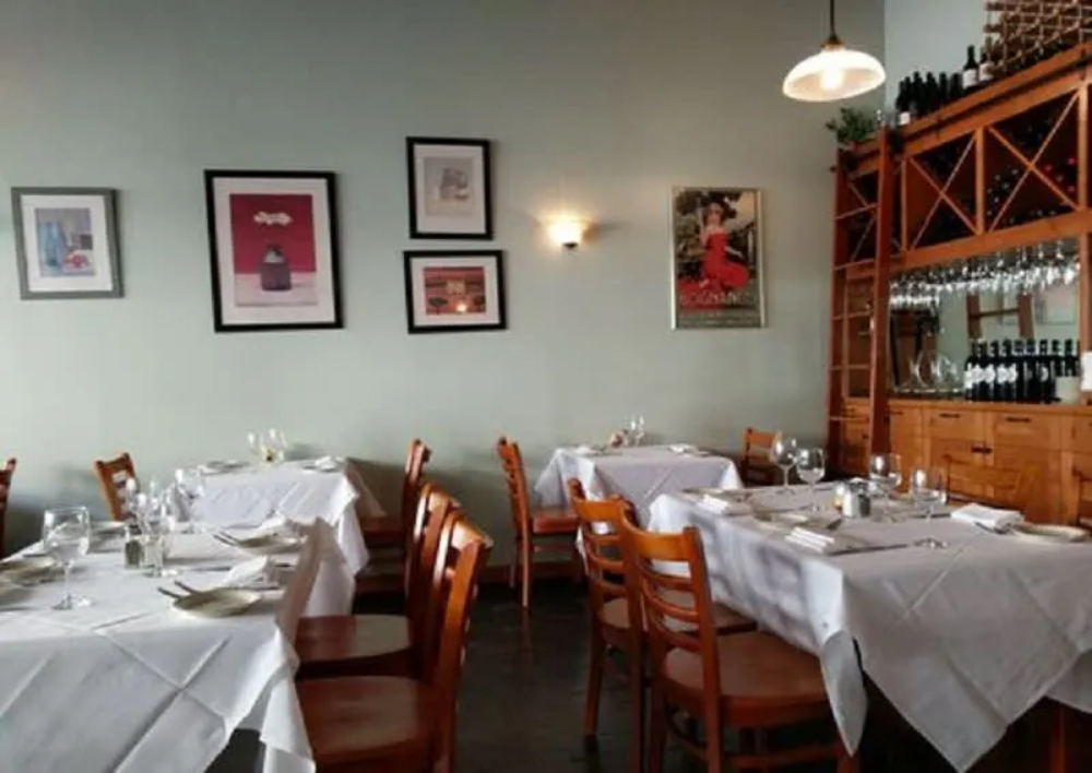 A cozy restaurant interior with wooden chairs, white tablecloths, framed artwork on the walls, and a wine display.