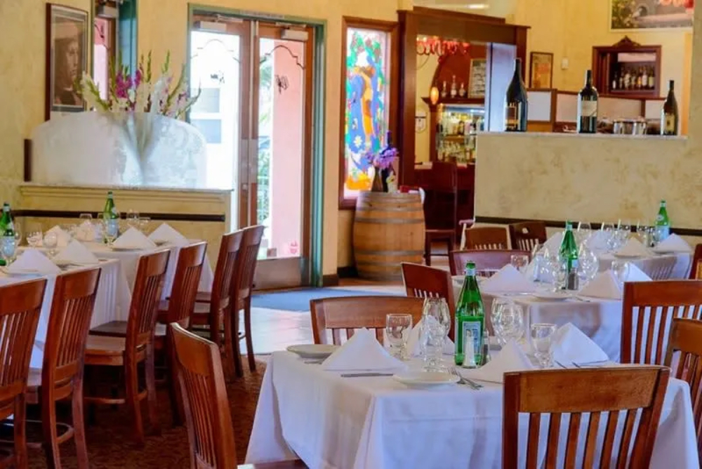 A cozy restaurant interior with wooden tables, white tablecloths, and decorative glass windows. A bar area is visible in the background.