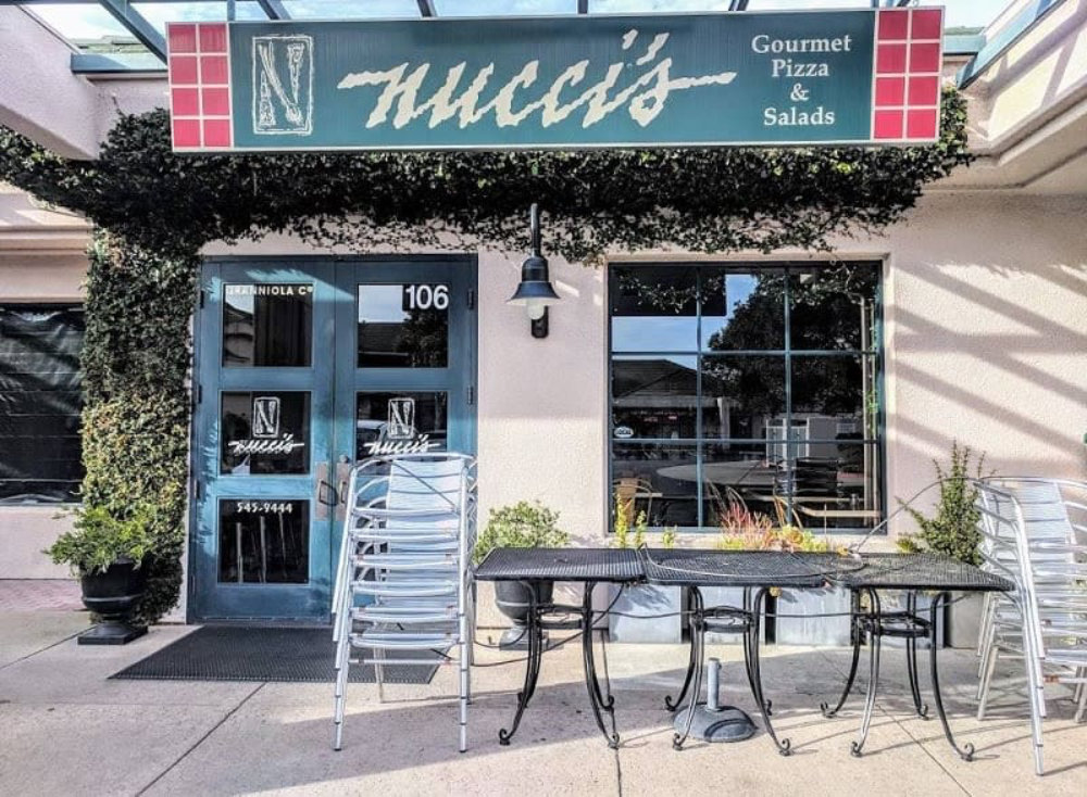 Exterior view of Nucci's restaurant featuring a sign for gourmet pizza and salads, with outdoor seating and potted plants.