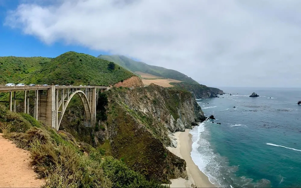 A scenic view of a coastal bridge spanning a cliffside, with lush green hills and a calm ocean below.