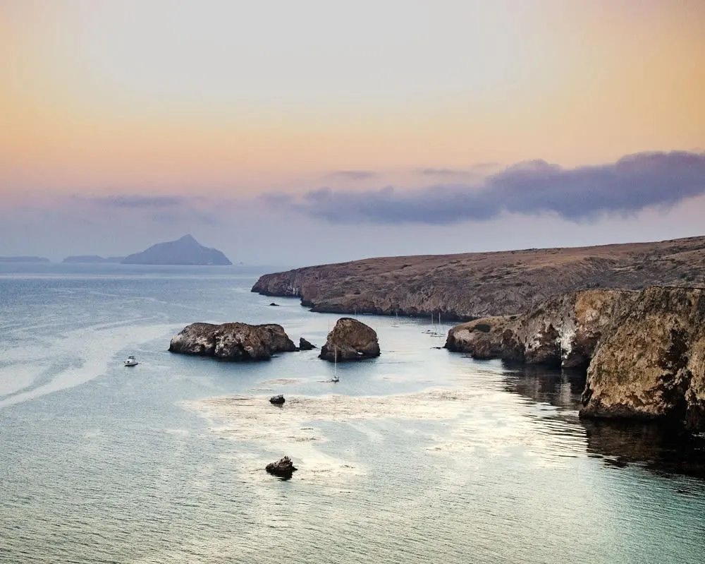 A serene coastal view at dusk, featuring rocky formations, calm waters, and distant islands under a colorful sky.