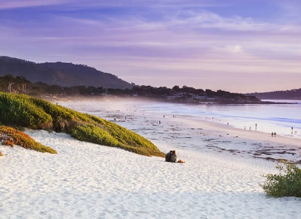 A serene beach scene at sunset, featuring gentle waves, sandy shores, and people walking along the coastline.