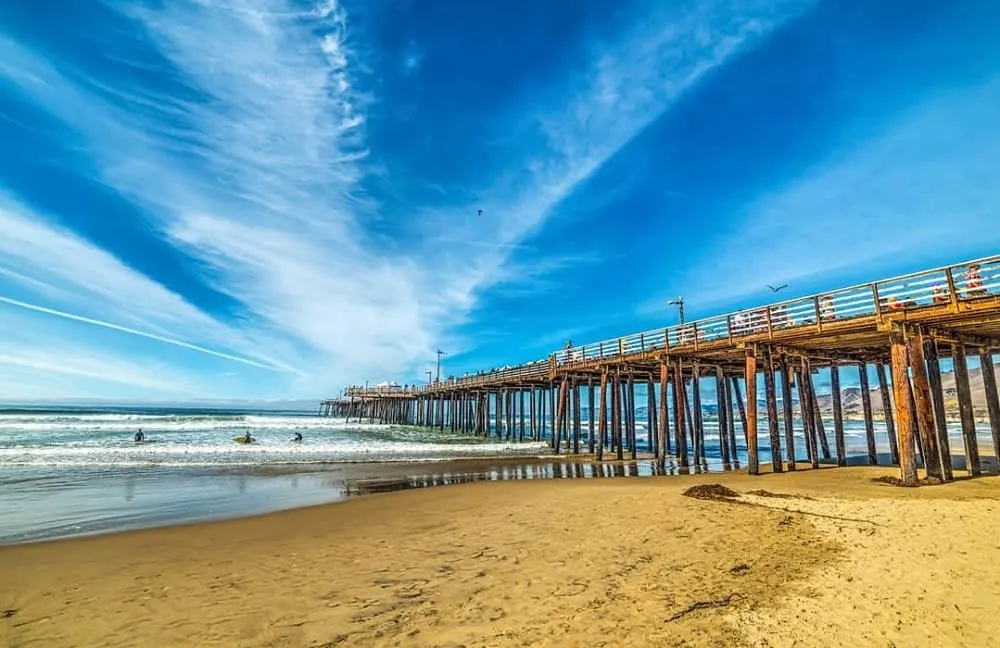 A wooden pier extends over a sandy beach, with surfers in the waves and a bright blue sky filled with wispy clouds.