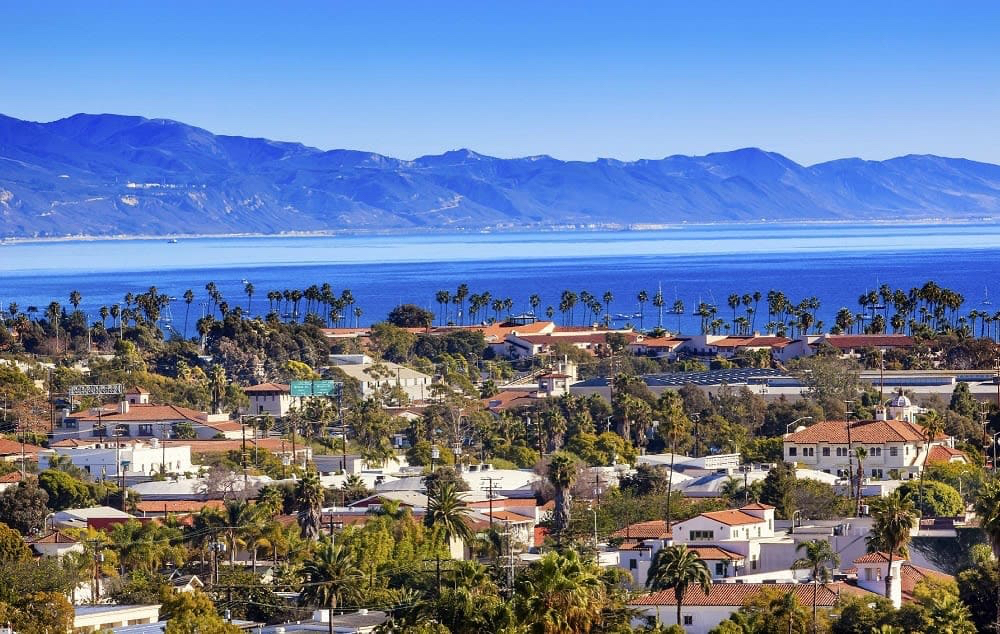 A scenic view of a coastal town with palm trees, houses, and mountains in the background under a clear blue sky.