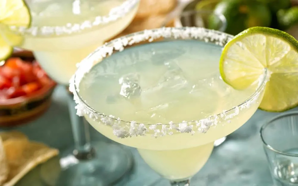 Two margaritas with salt rims and lime slices, served over ice, with chips and fresh ingredients in the background.