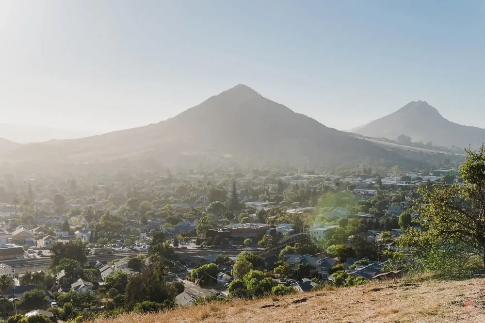 A scenic view of a valley with houses, surrounded by mountains under a hazy sky. Sunlight creates a warm atmosphere.