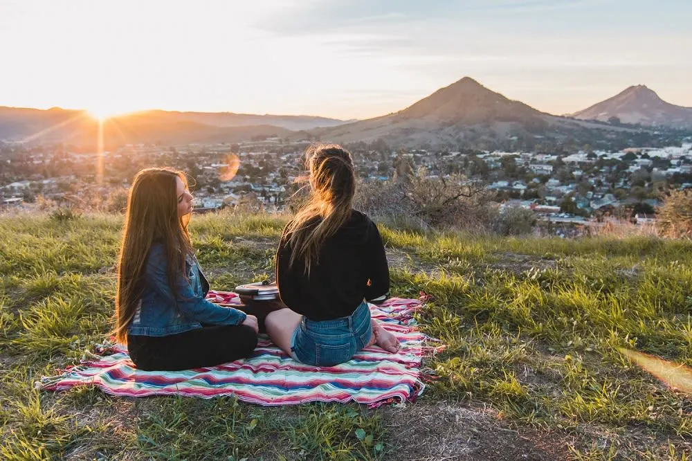 Two friends sit on a blanket, enjoying a sunset view over a town and mountains in the background.