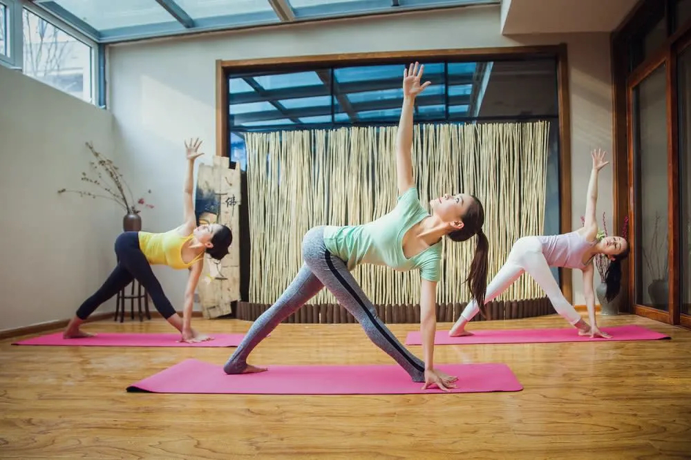 Three women practicing yoga in a bright studio, each in a different pose on pink mats. Natural light filters through large windows.