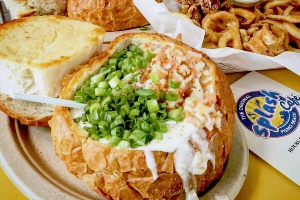 A bread bowl filled with creamy soup and topped with green onions, accompanied by toasted bread and side dishes.