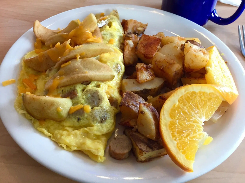 A plate featuring an omelette topped with apple slices and cheese, alongside seasoned potatoes and orange slices.