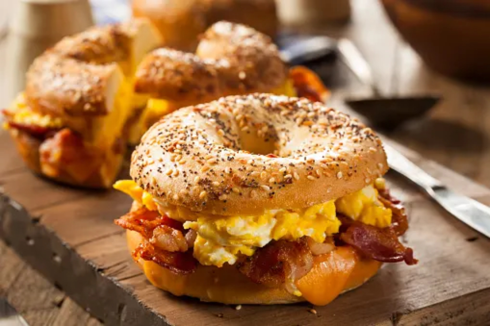 A toasted bagel sandwich filled with scrambled eggs and crispy bacon, served on a wooden board.