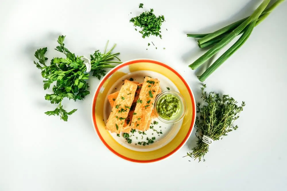A plate with seasoned fish, garnished with herbs, accompanied by a small bowl of green sauce and fresh herbs on the side.