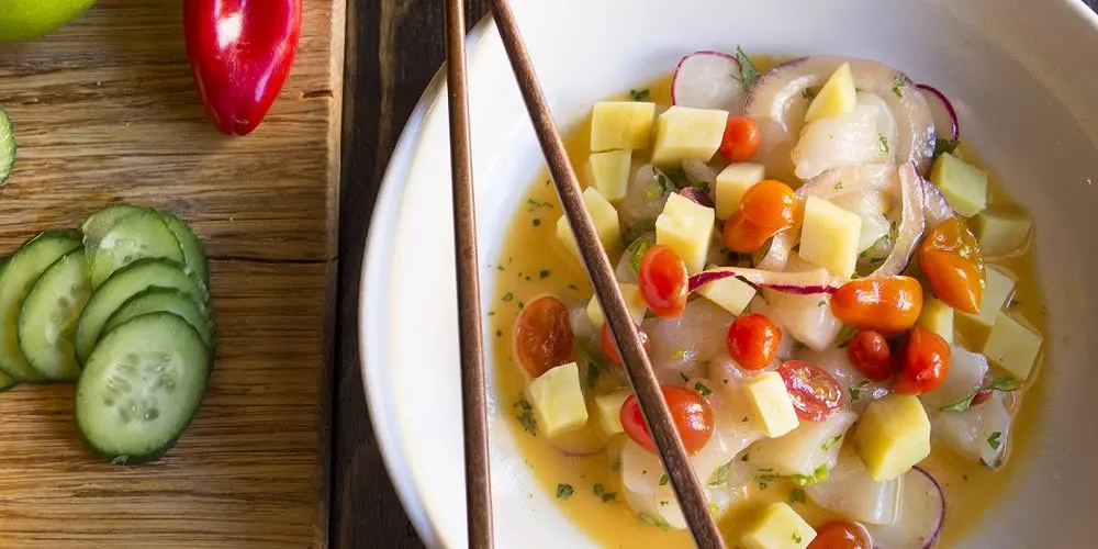 A bowl of ceviche with diced fish, mango, cherry tomatoes, and herbs, accompanied by sliced cucumbers and a red chili on a wooden surface.