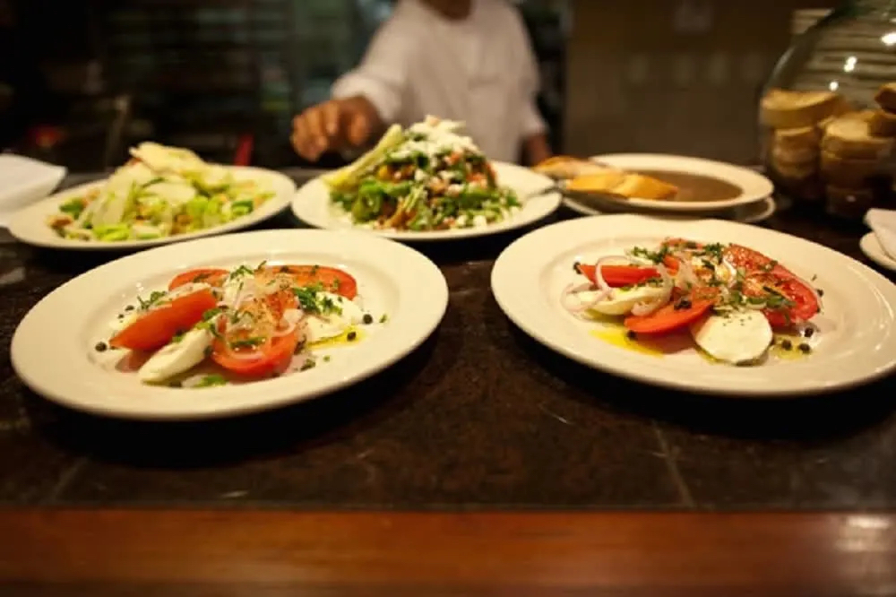 Three plates of fresh salad with tomatoes, mozzarella, and herbs, set on a countertop with additional dishes in the background.