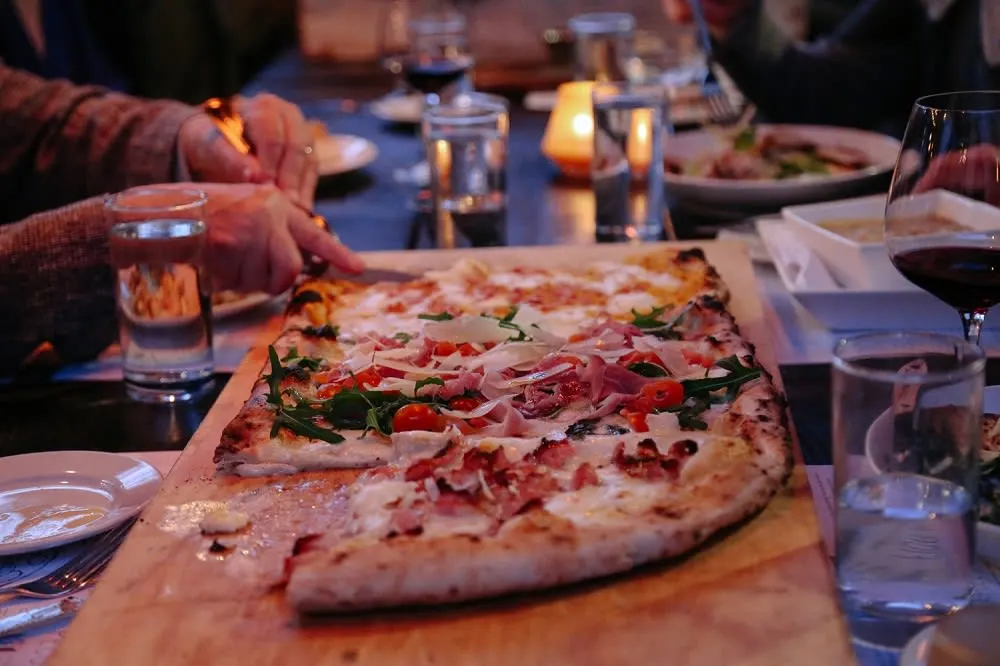 A wooden board with a large pizza topped with greens, tomatoes, and meats, surrounded by glasses of water and wine.