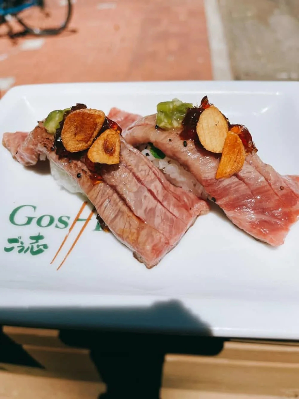 Two pieces of sushi topped with sliced garlic and green condiments on a white plate.