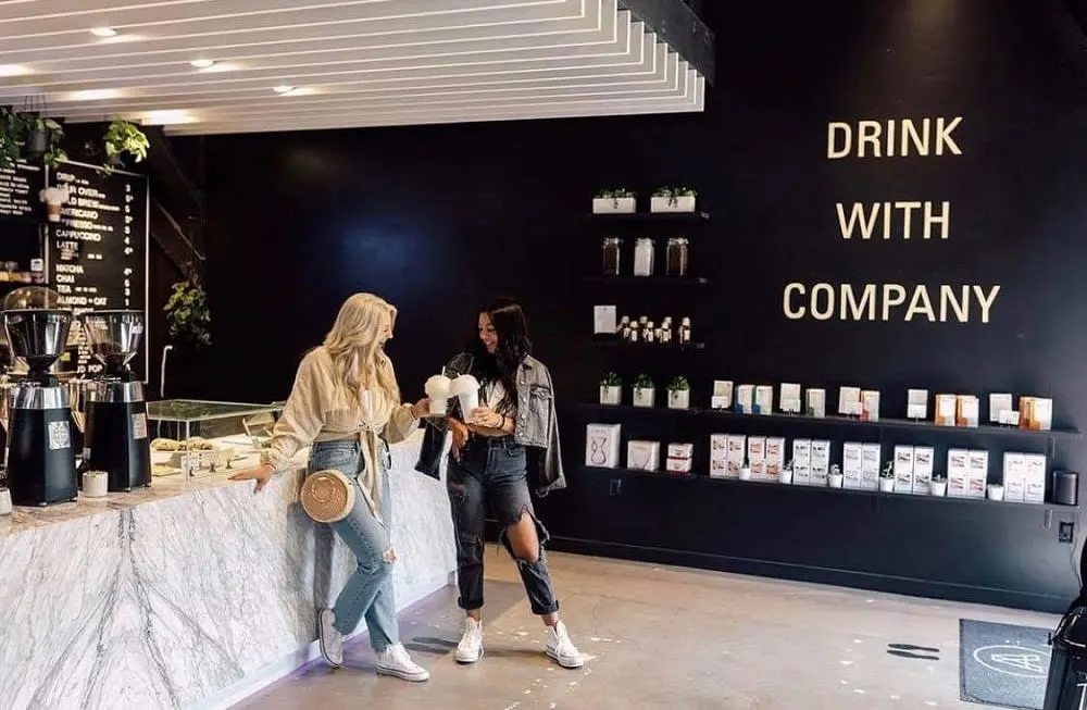 Two women chat at a stylish coffee shop with a marble counter and a black wall featuring the phrase "Drink with Company."