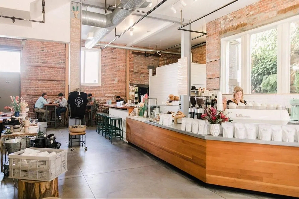 A cozy café with exposed brick walls, wooden counters, and a coffee bar. Customers are seated at tables, enjoying their drinks.