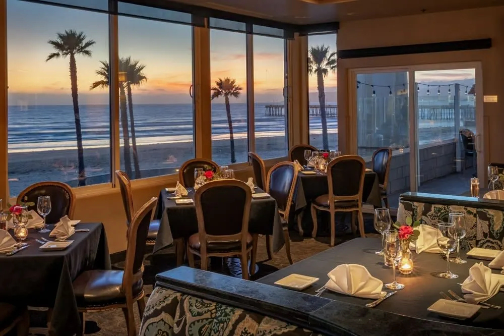A cozy restaurant with elegant tables, overlooking a beach at sunset, framed by palm trees and ocean views.