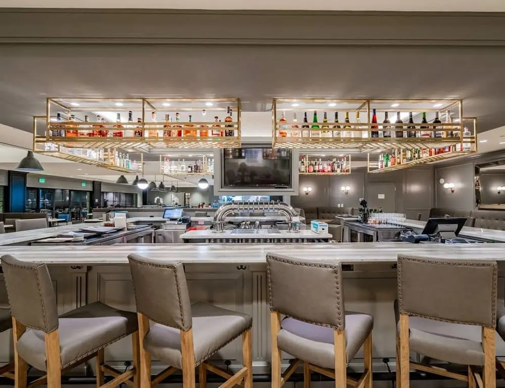 A modern bar with a sleek countertop, high stools, and a display of various liquor bottles overhead.