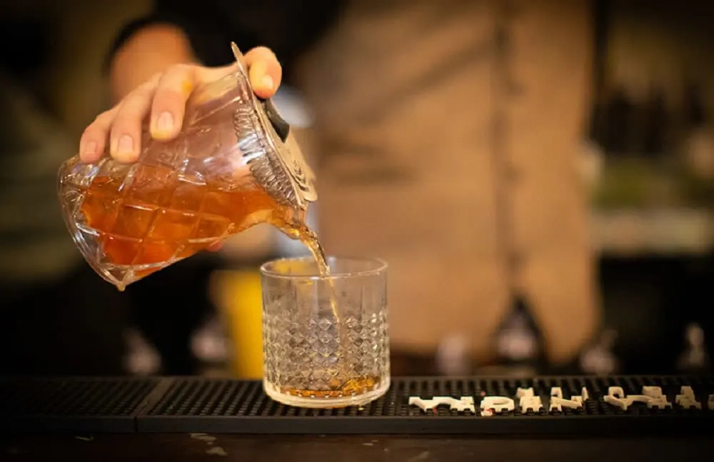 A bartender pours a brown liquid from a glass pitcher into a crystal glass on a bar counter.