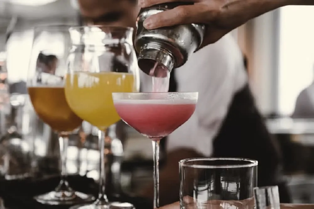 A hand pours a pink cocktail from a shaker into a glass, surrounded by other colorful drinks on a table.