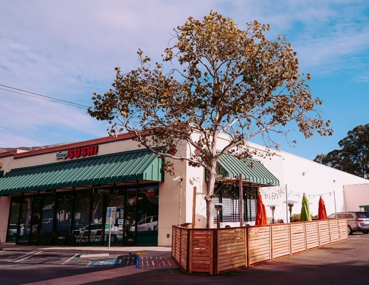 A sushi restaurant with a green awning, outdoor seating, and colorful umbrellas. A tree stands nearby under a blue sky.
