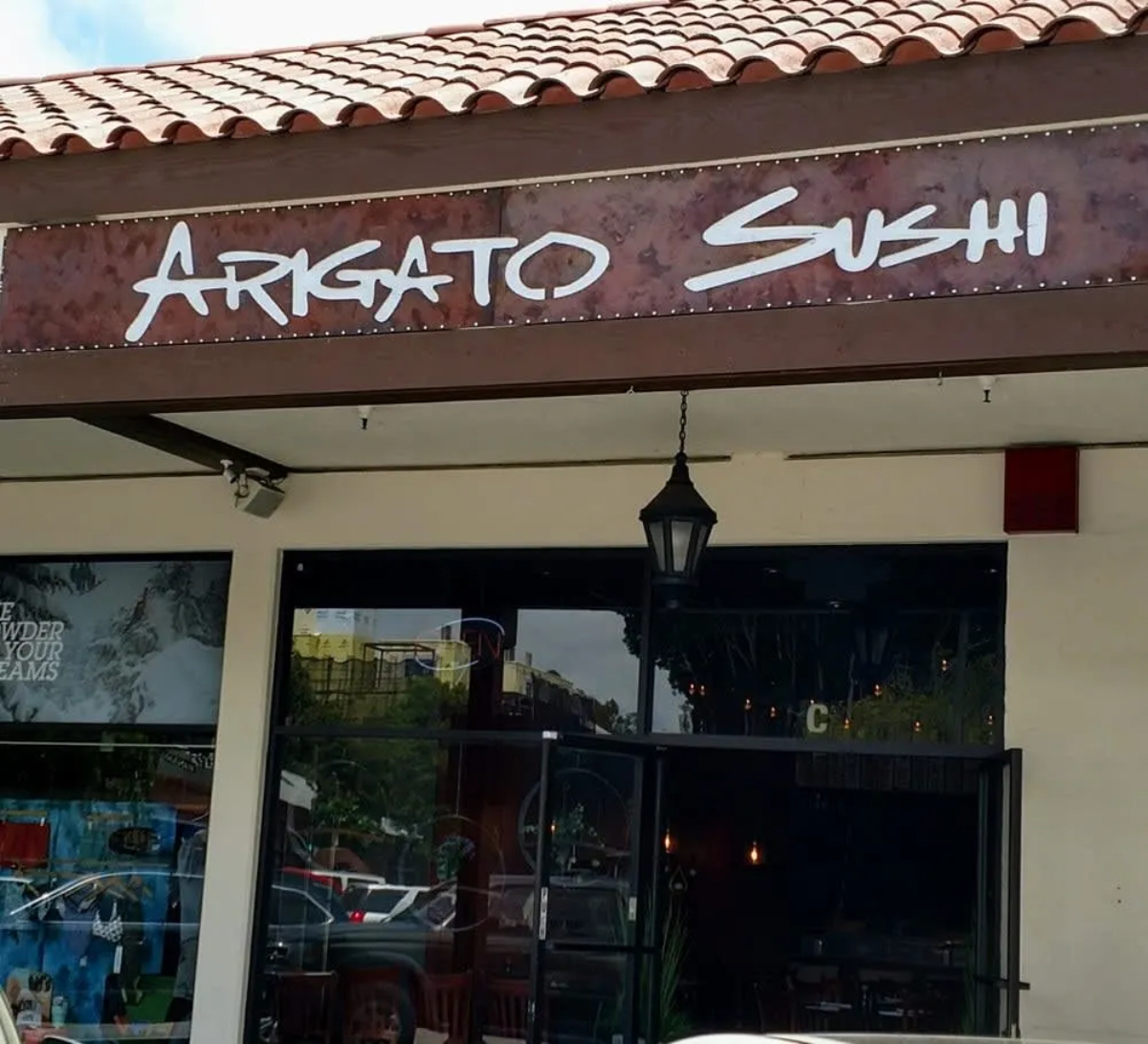 Sign for "Arigato Sushi" on a restaurant exterior with a tiled roof and a hanging lantern.