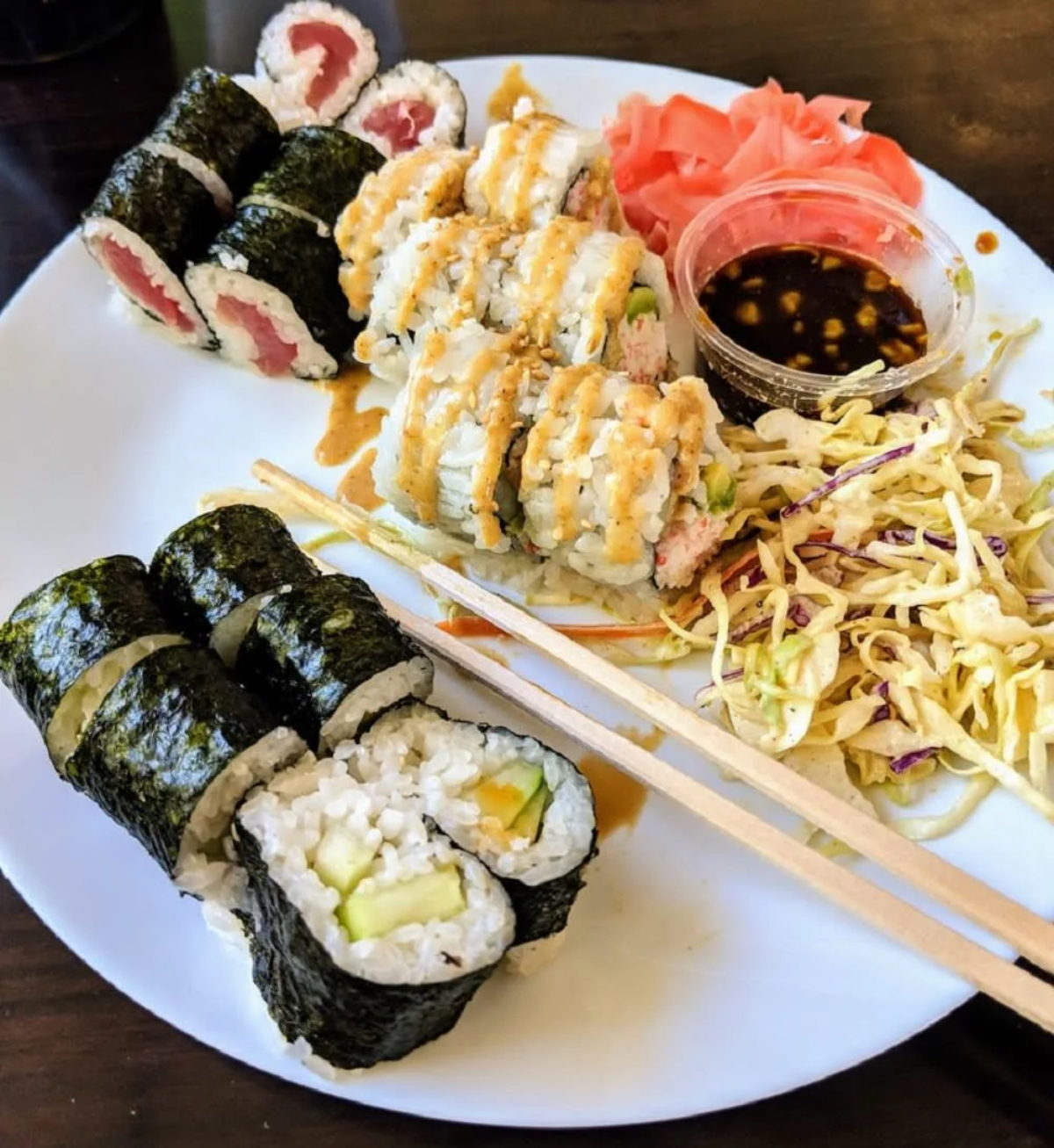 A plate of sushi featuring various rolls, pickled ginger, a small bowl of sauce, and a side of coleslaw.