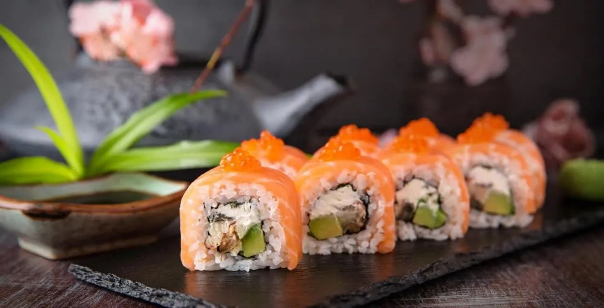A platter of sushi rolls topped with orange fish roe, served with a small dish of soy sauce and garnished with green leaves.