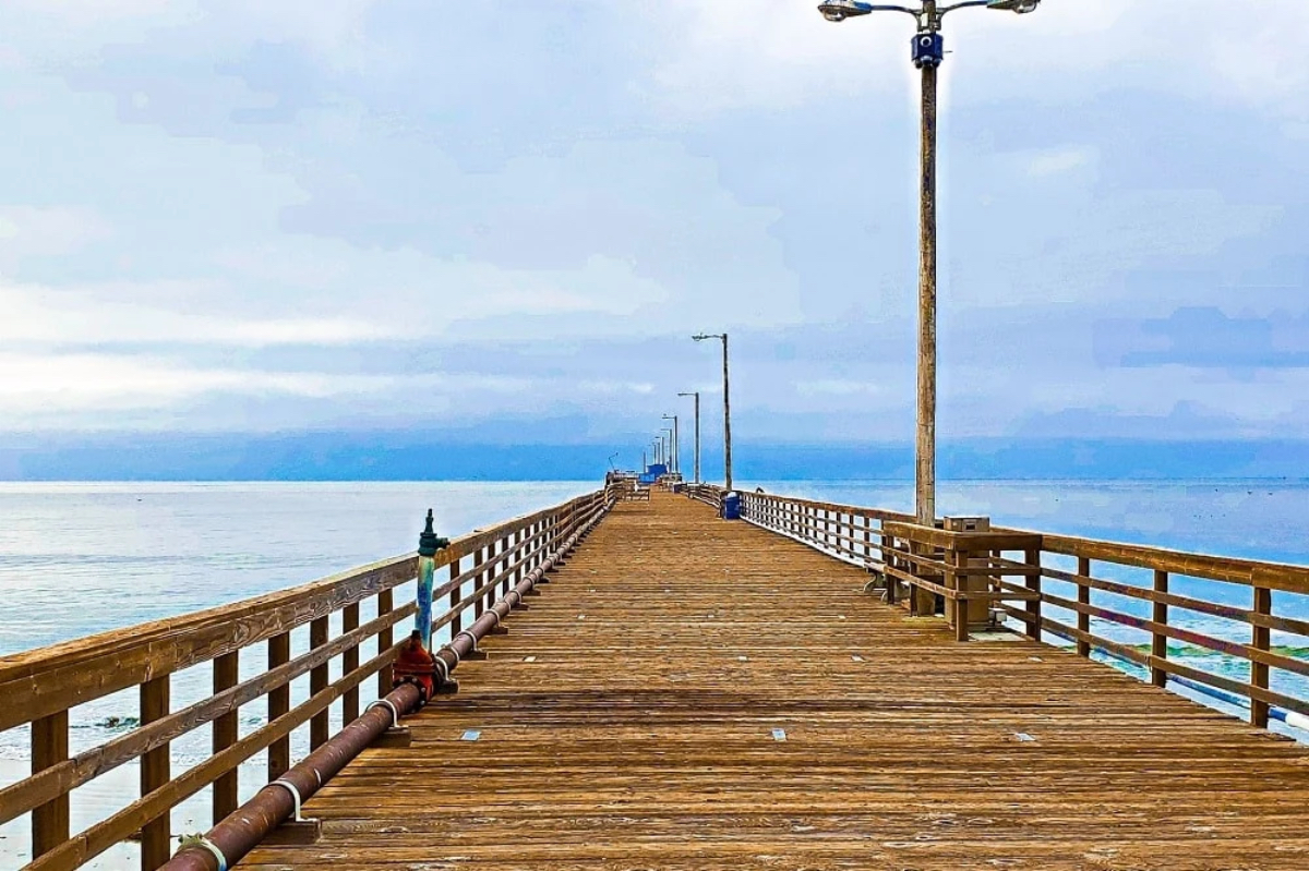 A wooden pier extends into calm waters, lined with light poles under a cloudy sky.