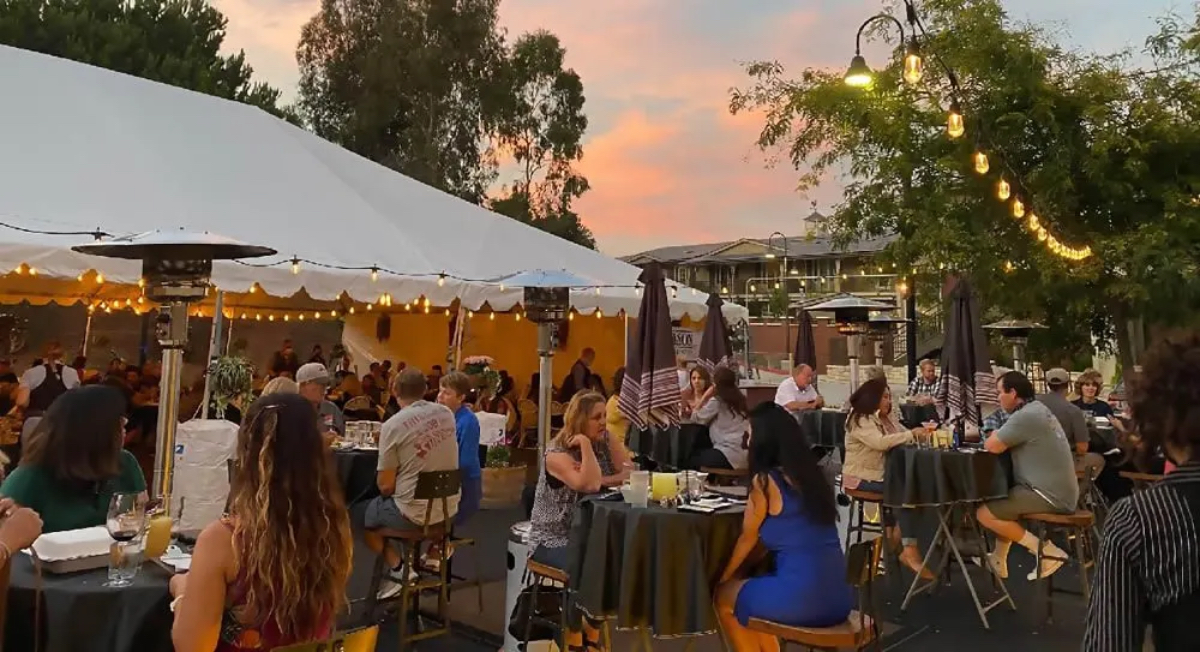 An outdoor dining scene under a large tent, with guests seated at tables, enjoying a sunset ambiance and string lights.
