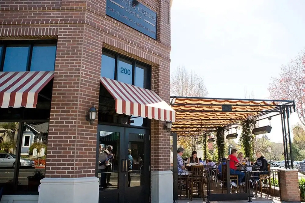 A brick building with striped awnings, featuring outdoor seating where people are dining. Trees and a clear sky are visible.