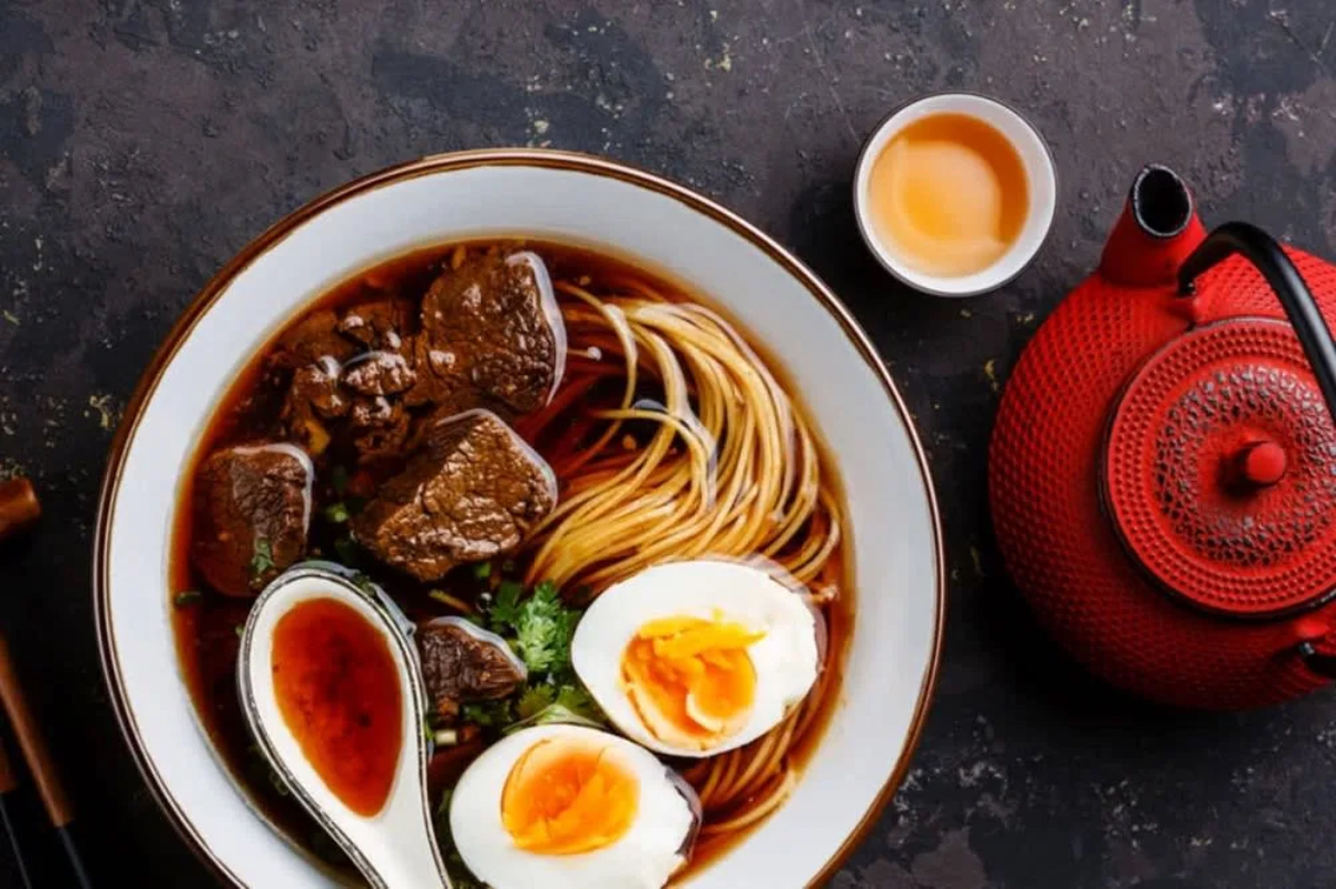 A bowl of beef noodle soup with soft-boiled eggs, herbs, and a side of tea, accompanied by a red teapot.