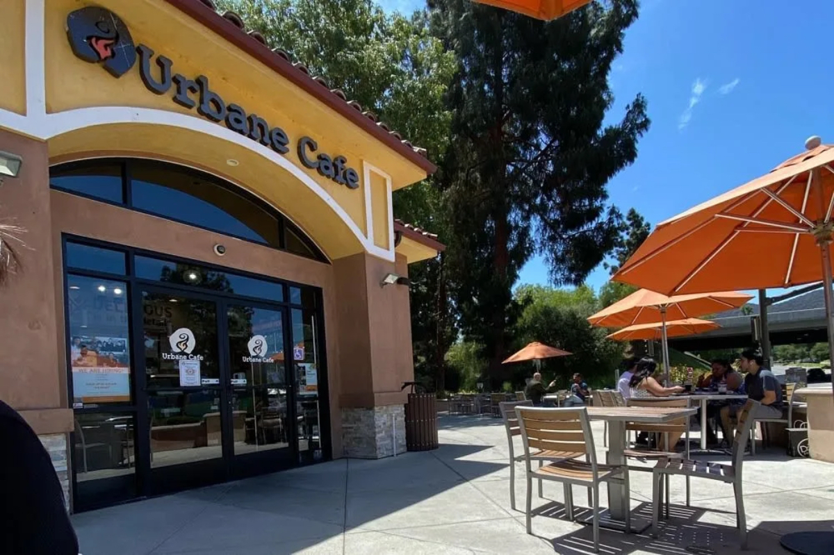 A sunny outdoor café scene featuring the Urbane Café entrance, with orange umbrellas and patrons seated at tables.
