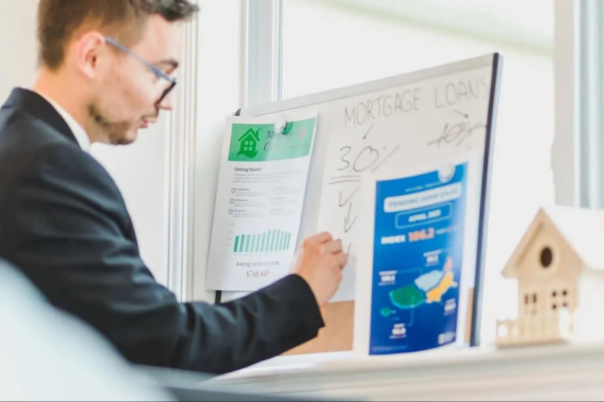 A man in a suit is reviewing mortgage loan information on a board, with charts and a small house model nearby.