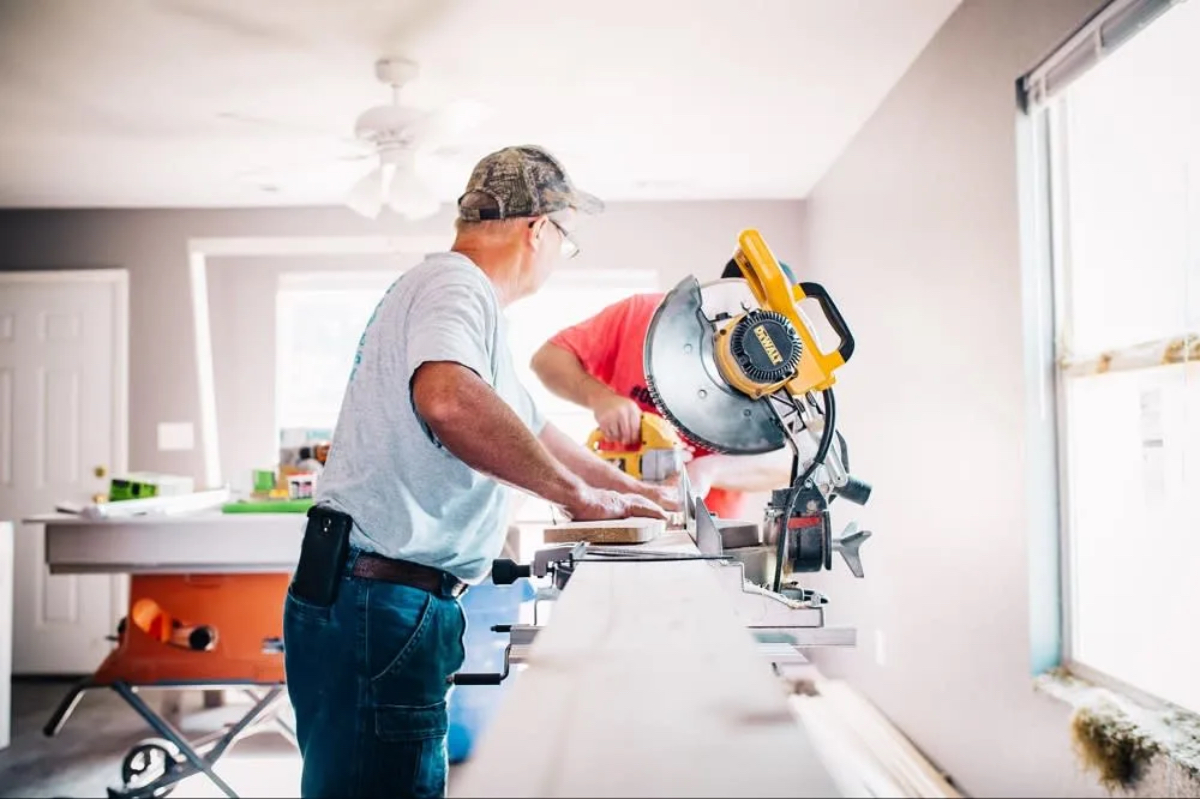 Two men are working on a woodworking project in a bright room, using a miter saw and handling wooden boards.