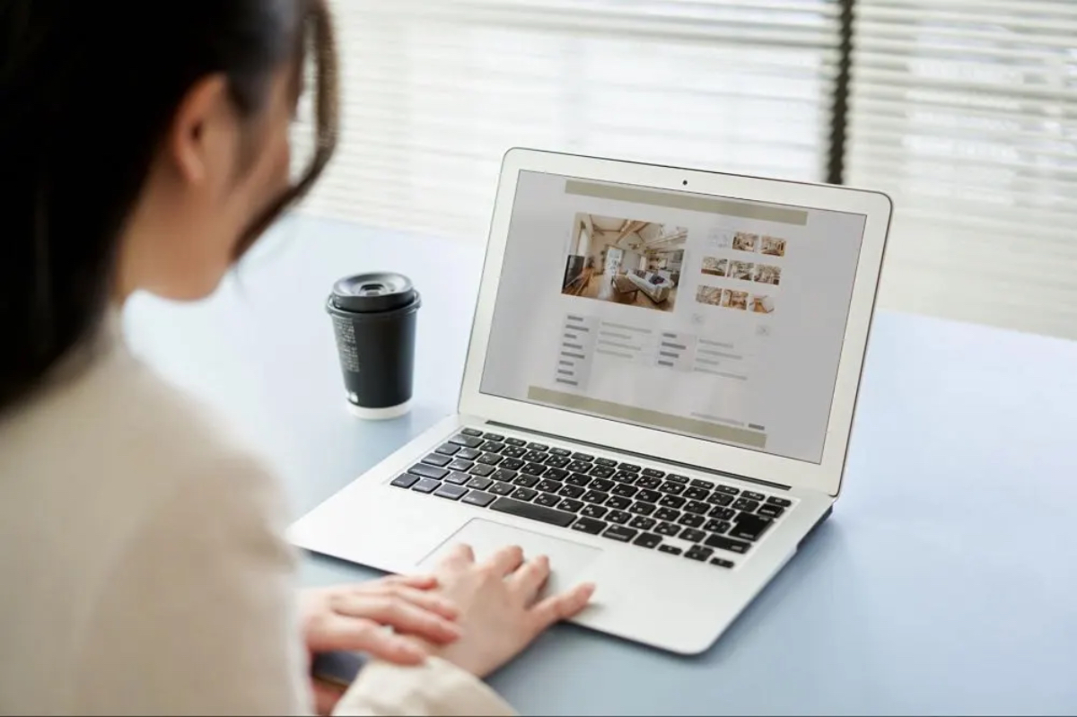 A person using a laptop at a table, viewing a webpage with images of interiors, alongside a coffee cup.